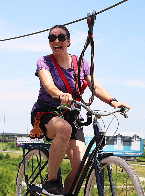a mom on a bike on a beam smiling as she crosses the ropes course